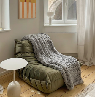 Cozy corner with a green cushion, gray blanket, and small white table in a room with large windows.